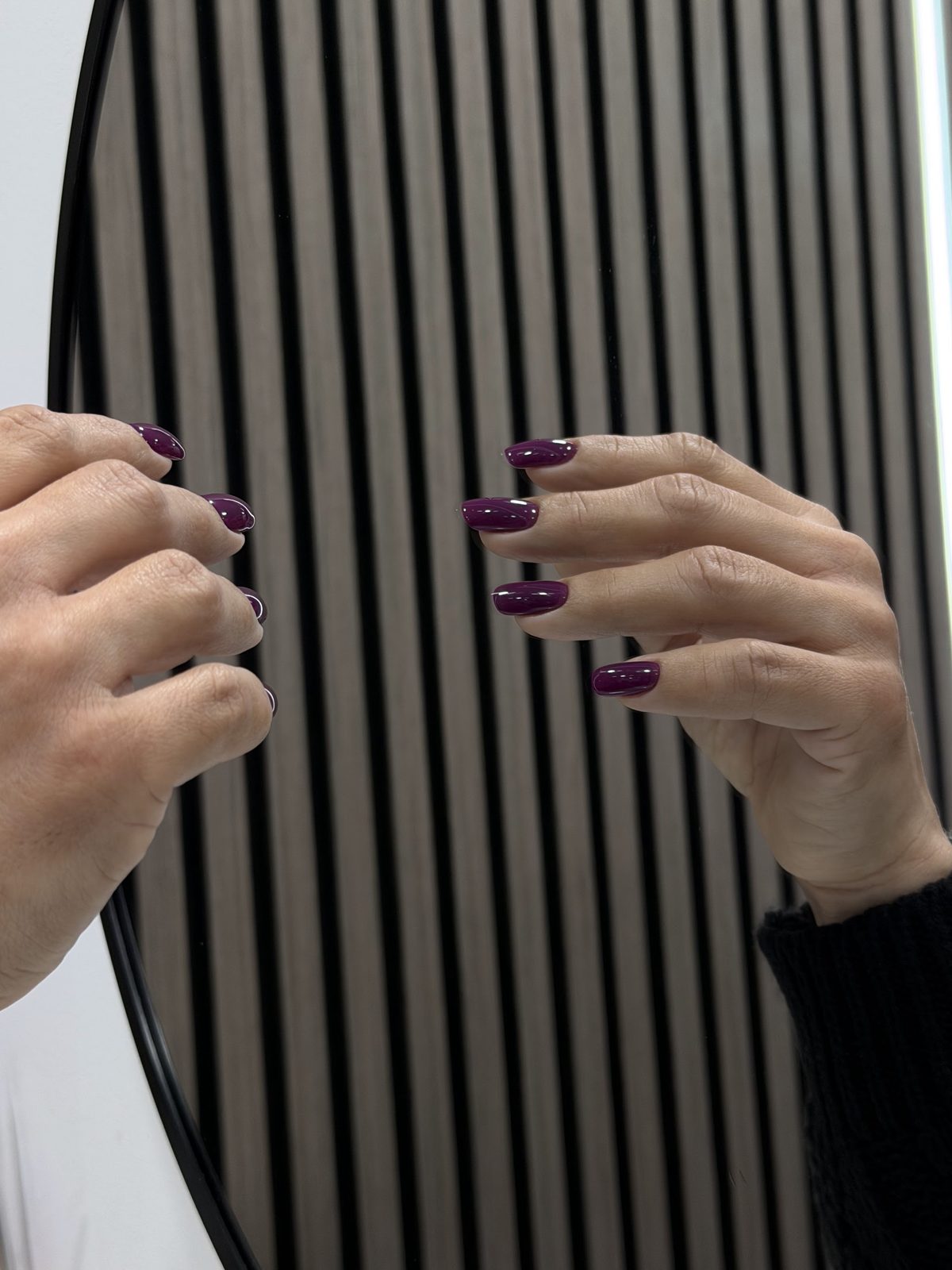 Twee handen met aubergine nagels voor de ronde spiegel en de houten lamellenwand van de salon